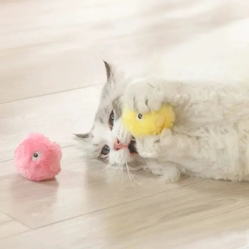 Two plush squeak toy balls for cats; yellow ball in paws, pink ball on floor.