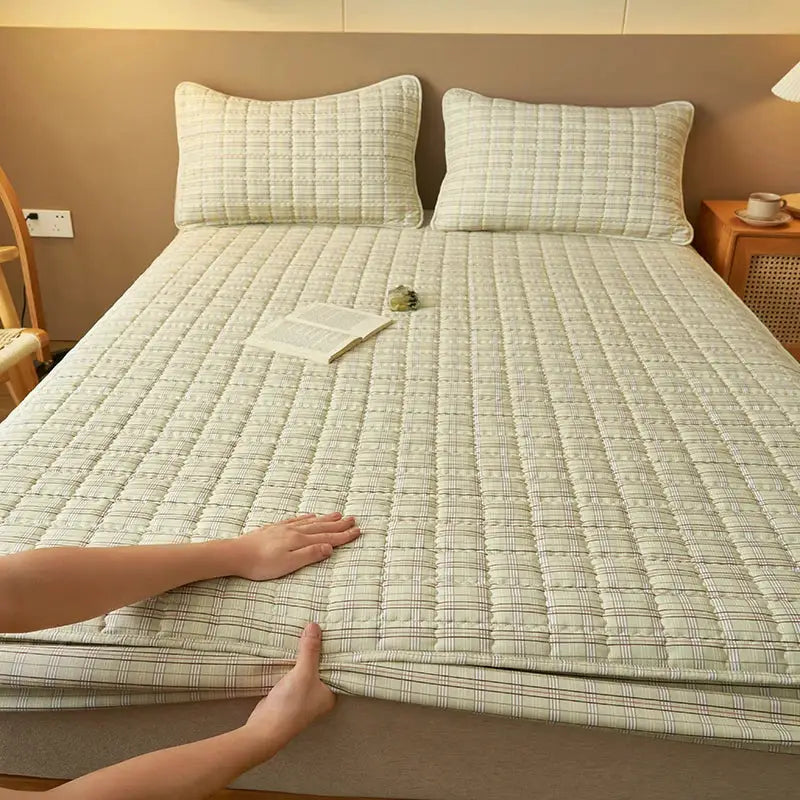 Quilted plaid mattress protector by Simples Market; antibacterial, breathable beige cover on a bed with two pillows.
