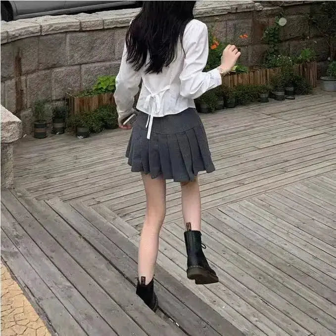 White women's blouse with back tie detail and long sleeves, paired with a gray pleated mini skirt and black ankle boots