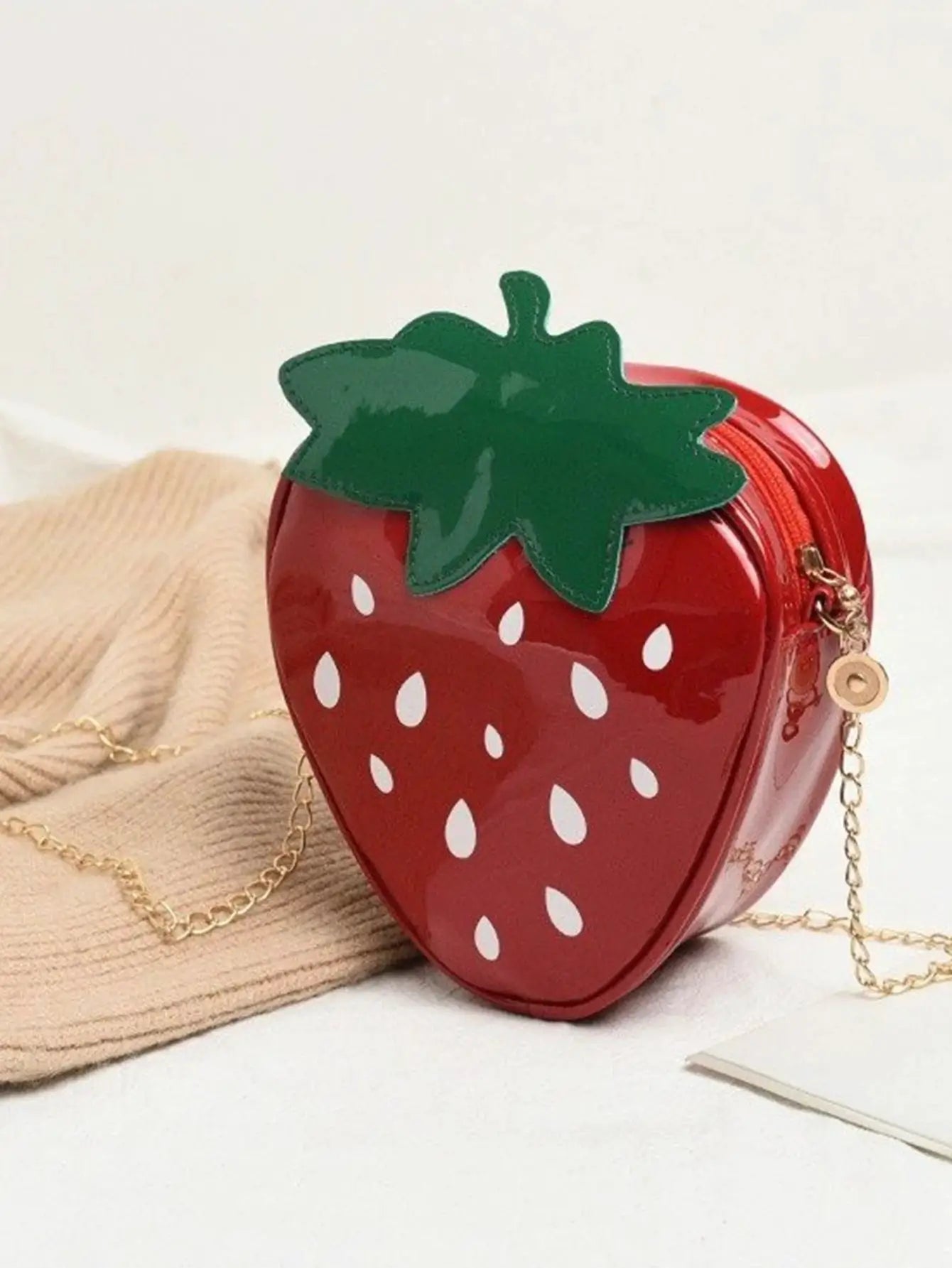 Strawberry-shaped crossbody bag in red with white seed accents, green leaf top, and gold chain strap.