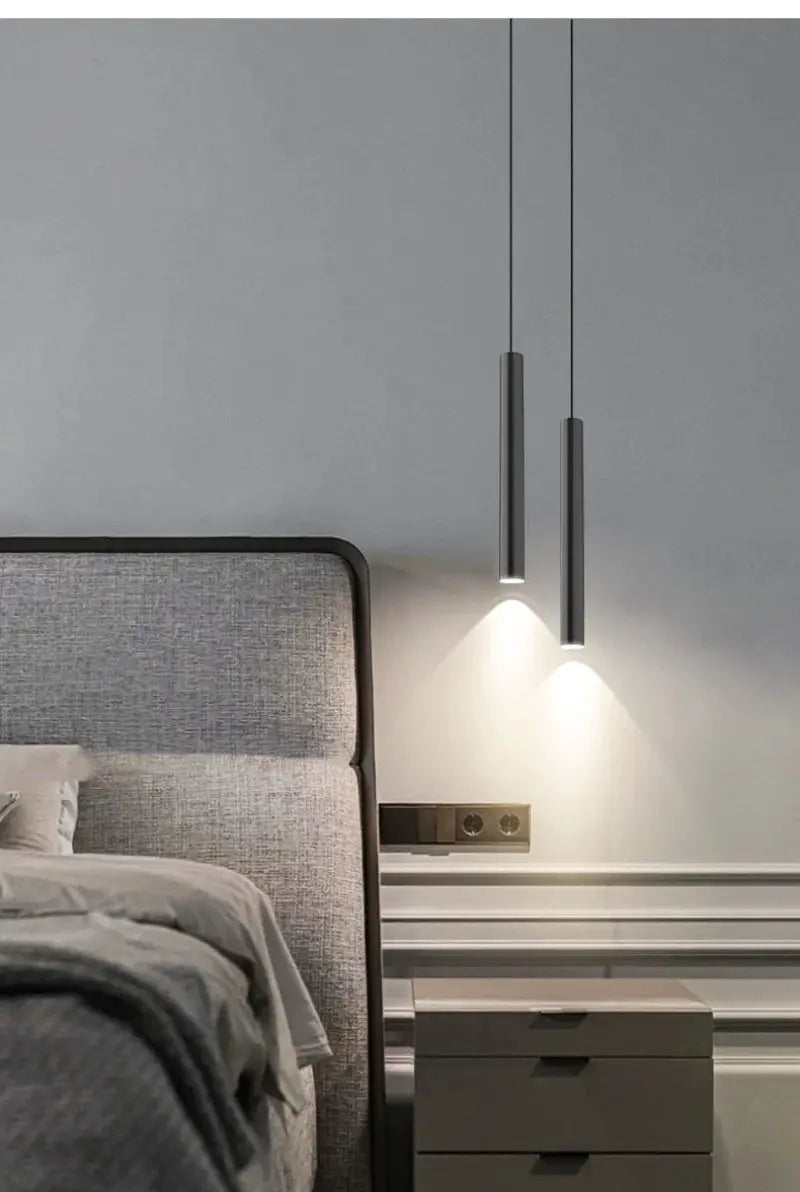 Minimalist bedside pendant lamps, two dark metal cylinders with LED downlights over a bedroom nightstand.