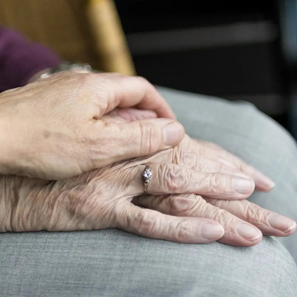 Two elderly hands folded on gray fabric, wearing a delicate ring.