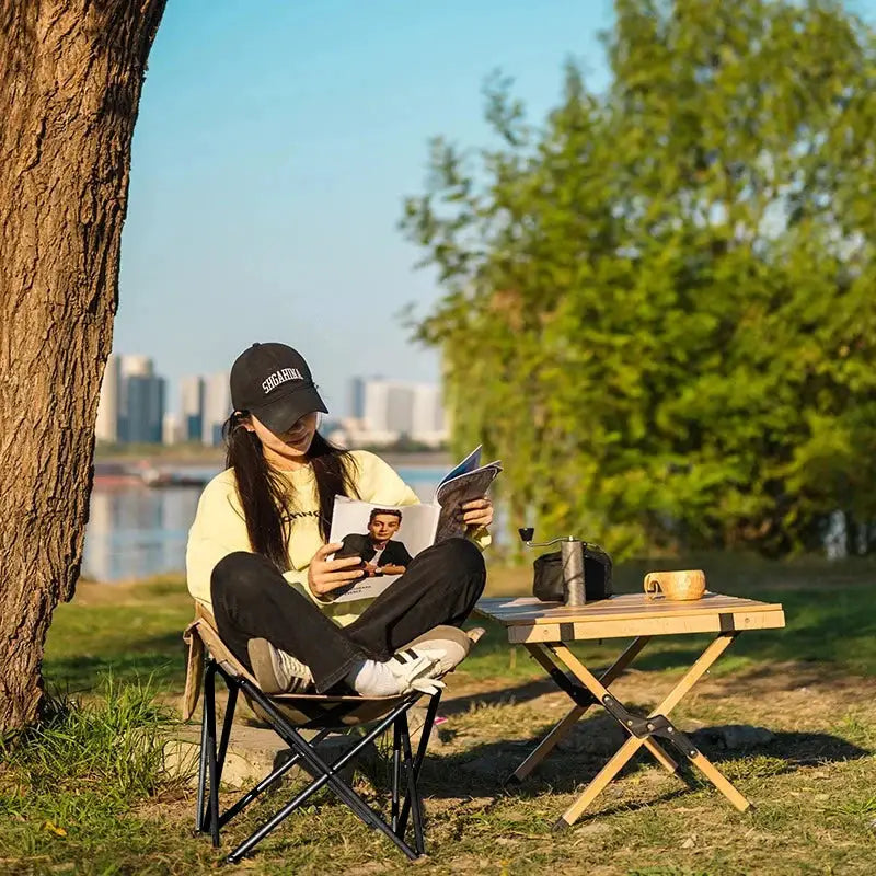 Outdoor folding moon chair with black frame and beige fabric seat beside a wooden folding picnic table.