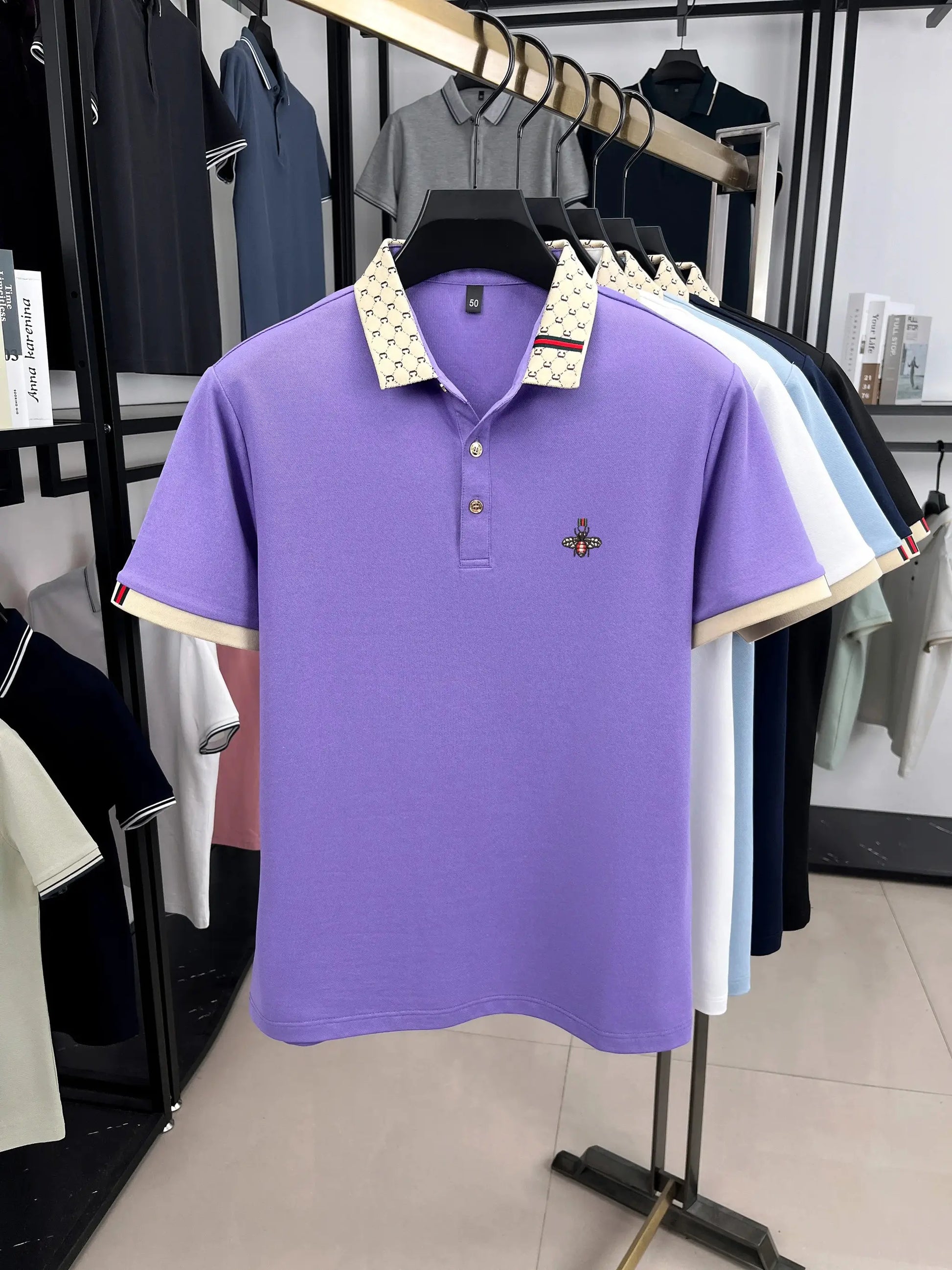 Polo shirt lavender with beige patterned collar and bee embroidery; five similar polos on display.