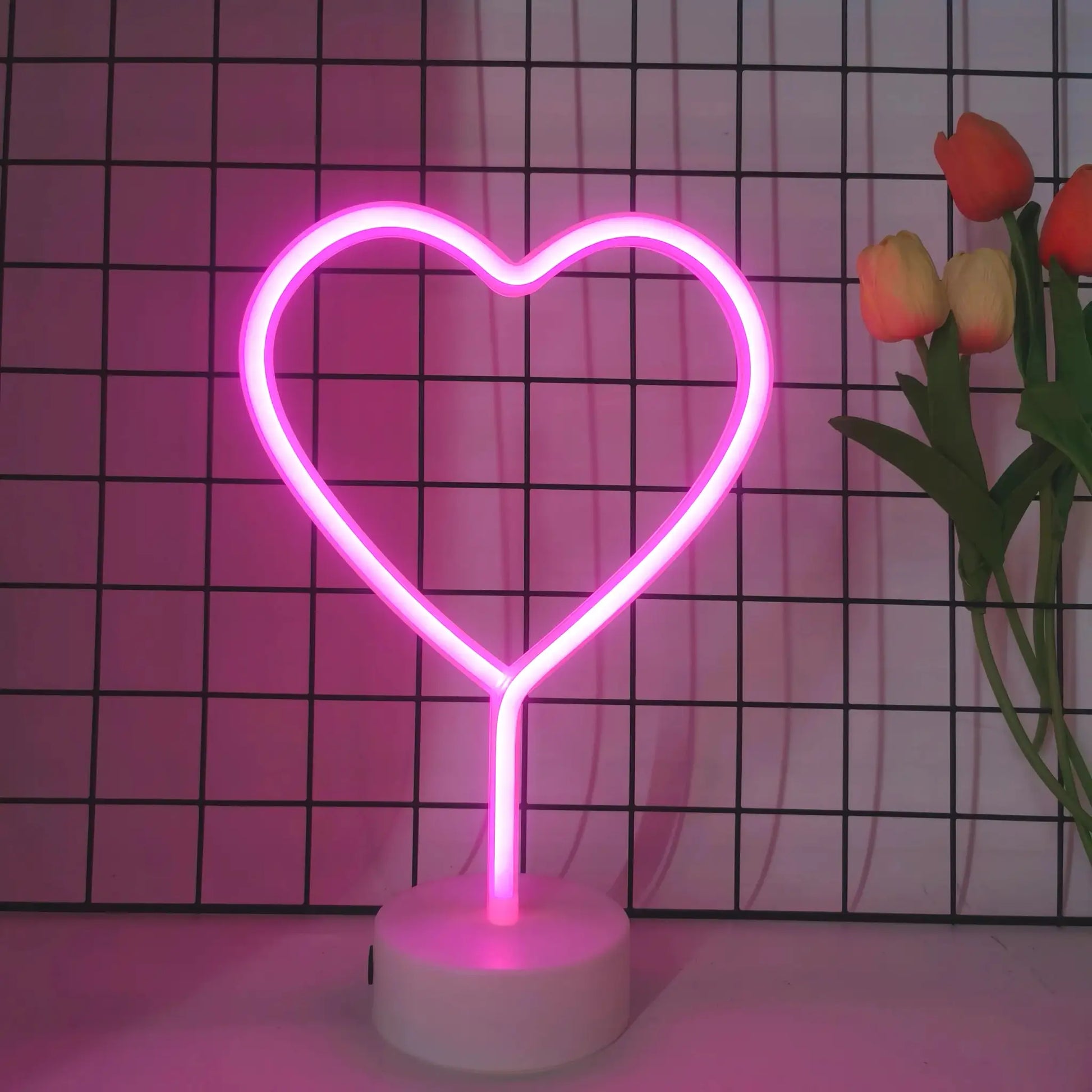 Pink heart-shaped neon light on a white base, USB or battery powered, with tulips in the background.