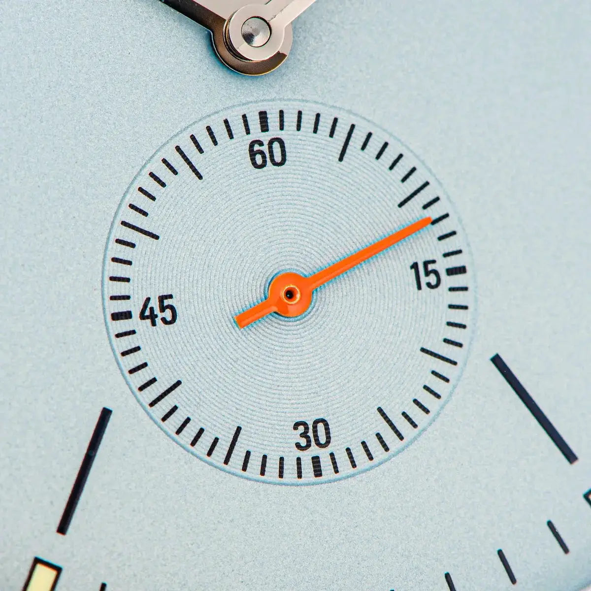 Men's wristwatch dial close-up with orange hands and bold black minute markers on a light blue background
