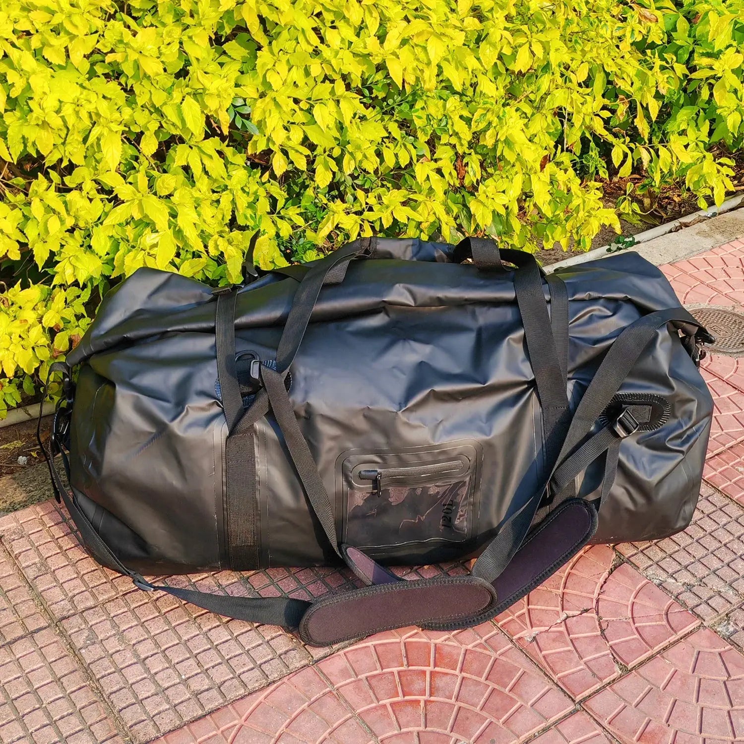 Black 40L-120L waterproof duffle bag with carry straps lying on pavement beside a yellow hedge