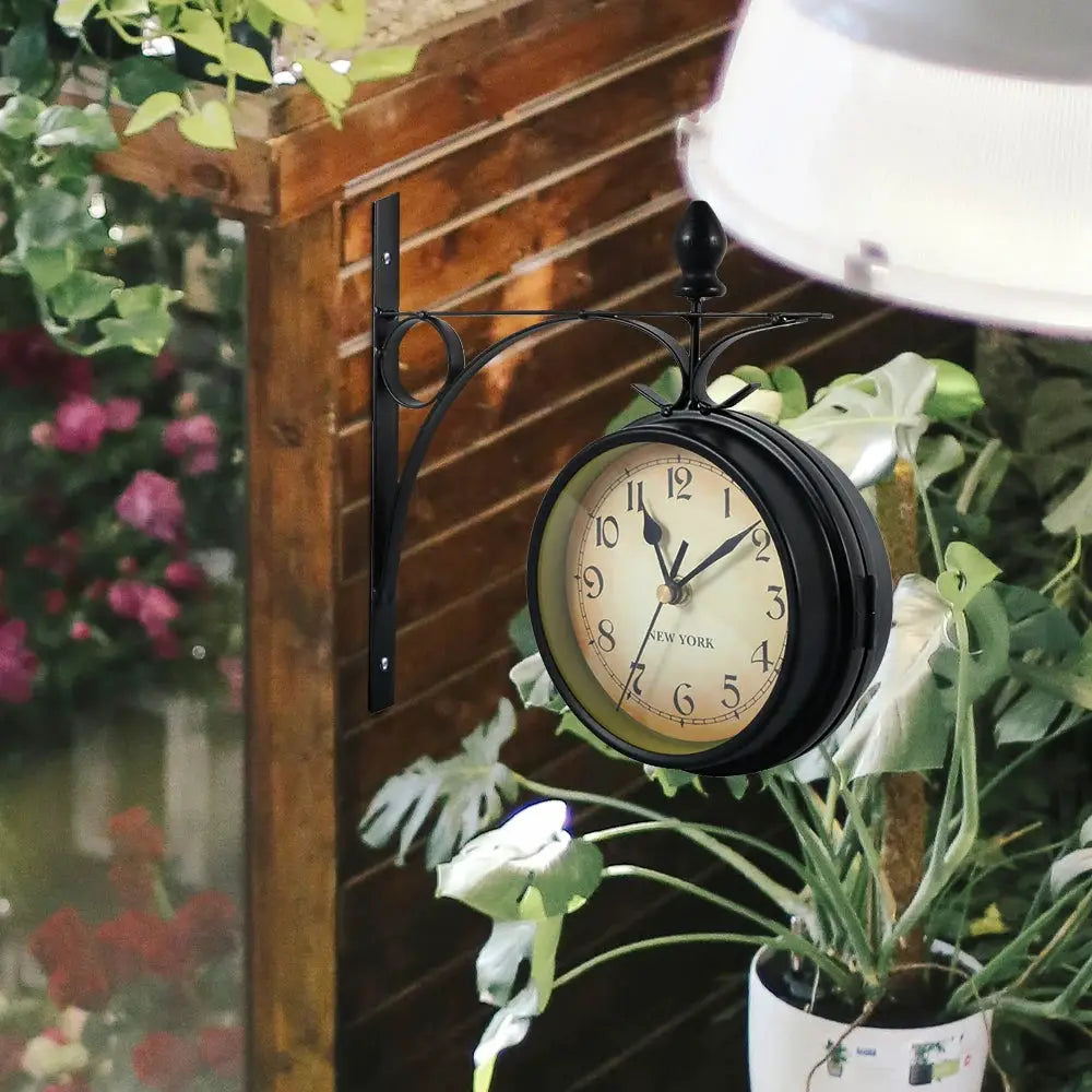 Wall clock mounted on a black decorative metal bracket with a round beige dial and bold black numerals, outdoor garden setting
