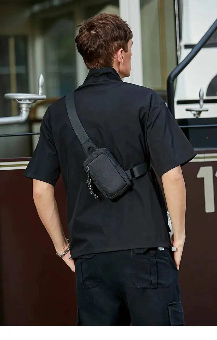 Black crossbody bag; compact mini chest bag with zip pocket worn diagonally across the torso.