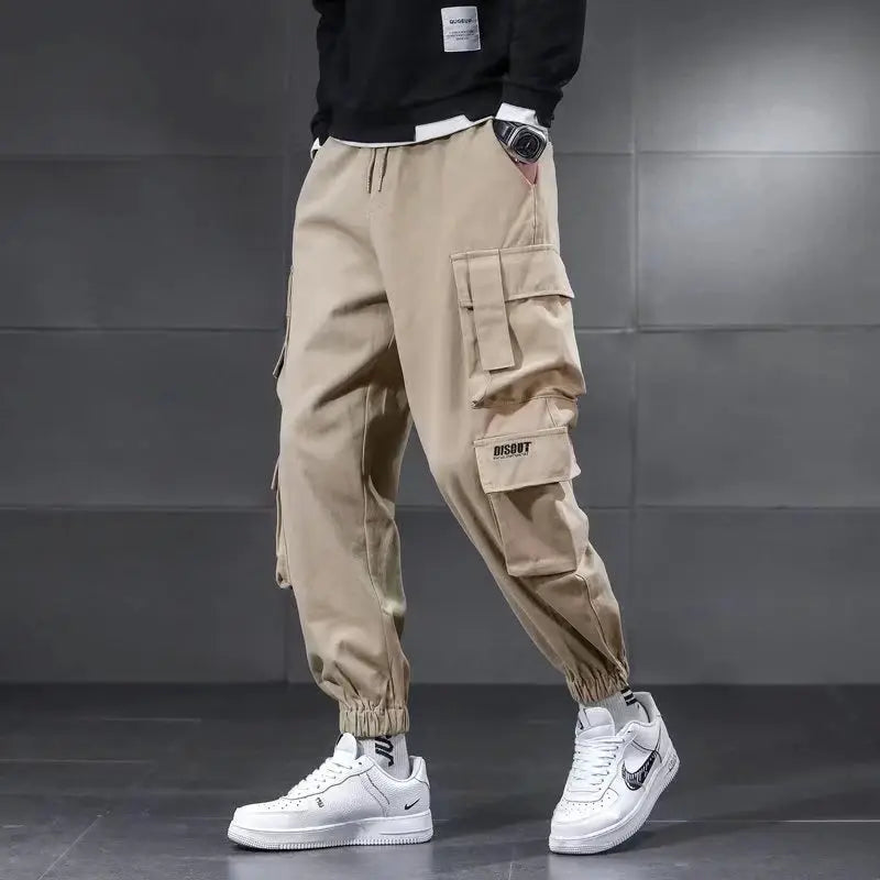 Men's beige cargo pants with DISOUT logo, multiple pockets, drawstring waist, and ribbed cuffs.