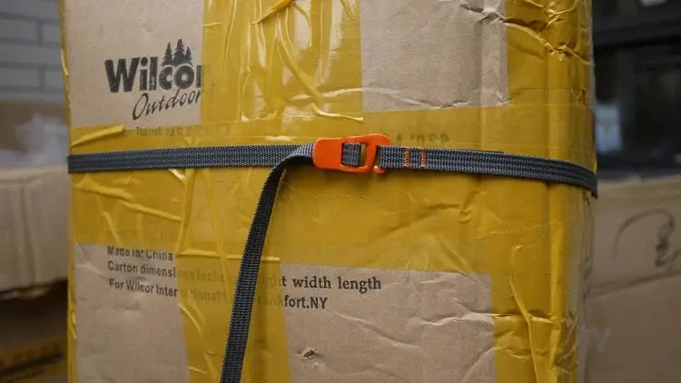 Luggage strap with orange quick-release buckle and gray nylon, wrapped around a yellow-taped cardboard box