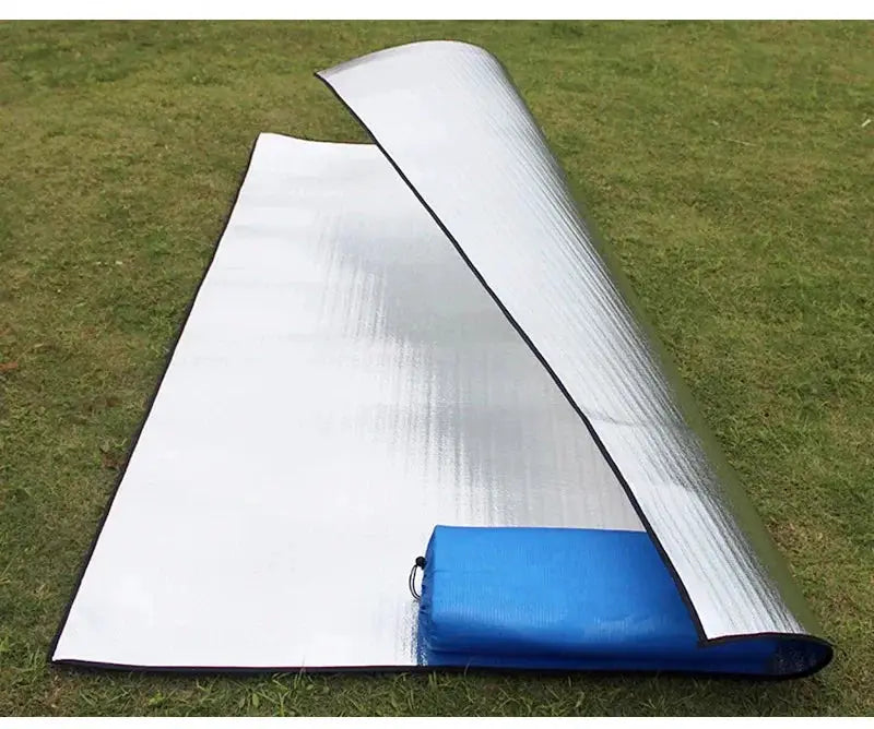 Camping mat with aluminum foil insulation; silver reflective sleeping pad and blue carry bag on grass.