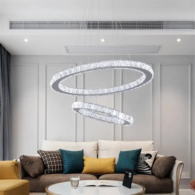 Modern crystal ring LED chandelier with two interlocking chrome rings, remote lighting over a living room sofa.