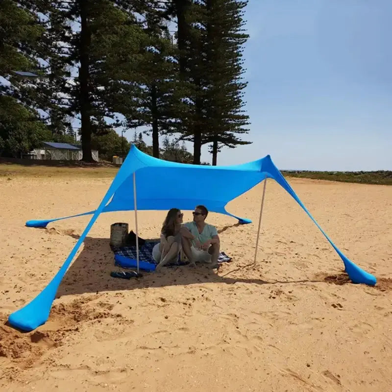 Blue windproof pop-up camping tent canopy on a sandy beach with two people sitting underneath