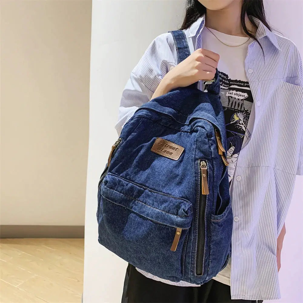 Denim backpack by Simples Market in blue with multiple pockets, large capacity, brown zipper pulls, and front pocket.