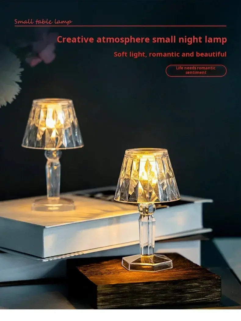 Pair of mini crystal table lamps with faceted shades and clear stems, battery-powered warm LED lights on stacked books and a wooden block