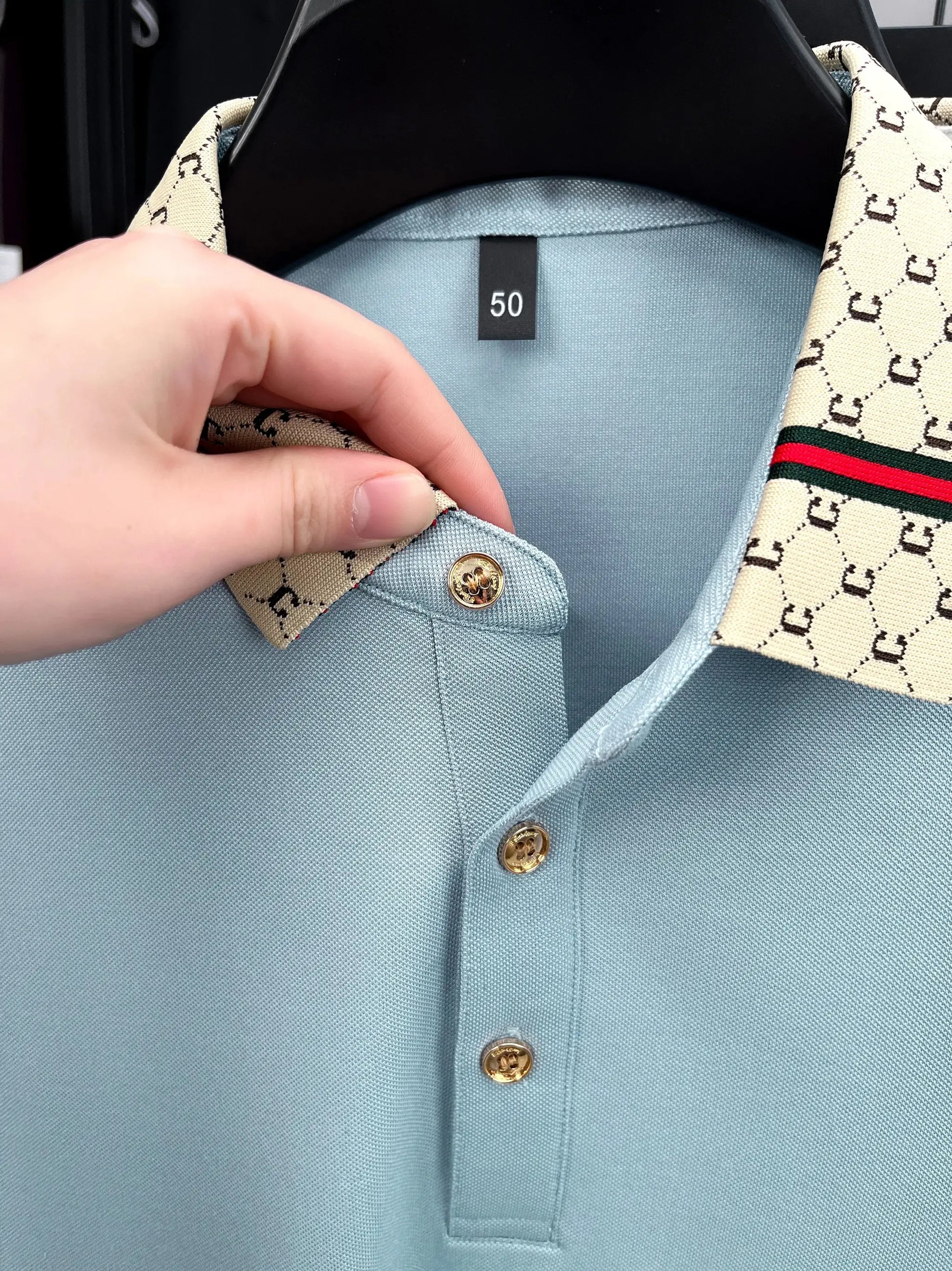 Men's polo shirt in light blue with beige monogram collar, red-green stripe, and gold buttons.
