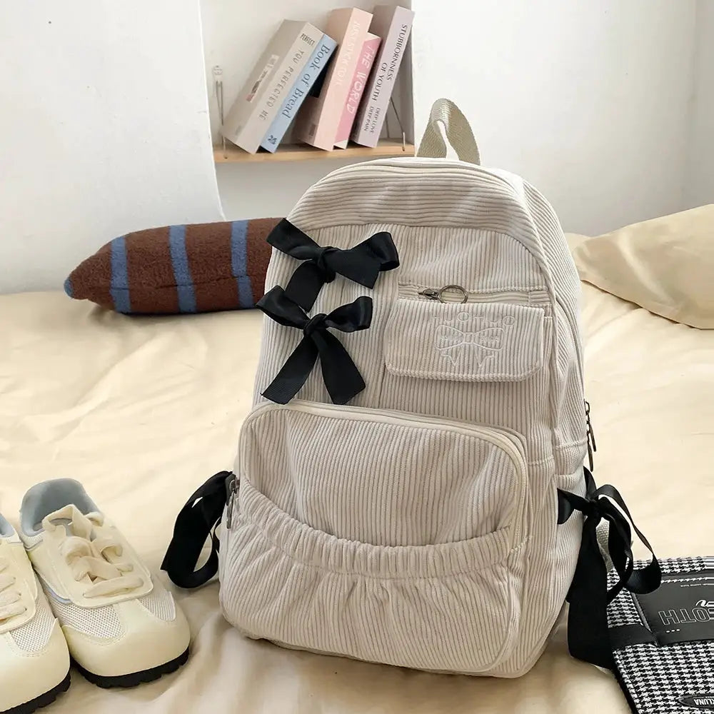 Beige corduroy backpack with two black bows, front zip pocket, and top handle.