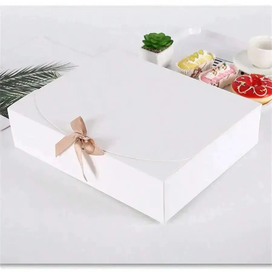 White rectangular gift box with lid and beige ribbon, displayed on a white surface beside desserts.