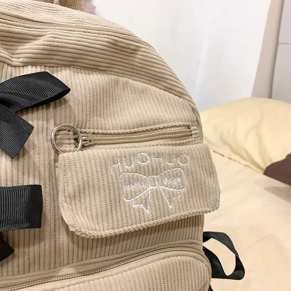 Backpack in beige corduroy with black straps and front bow embroidery, metal zippers.
