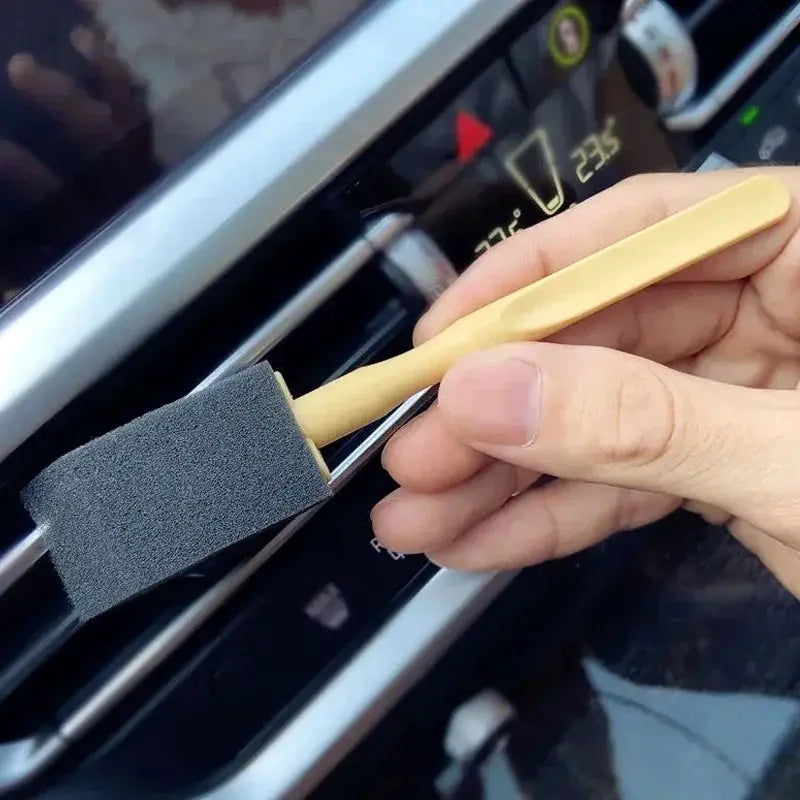Car AC vent cleaning brush with beige wooden handle and gray foam sponge being used on a car interior vent