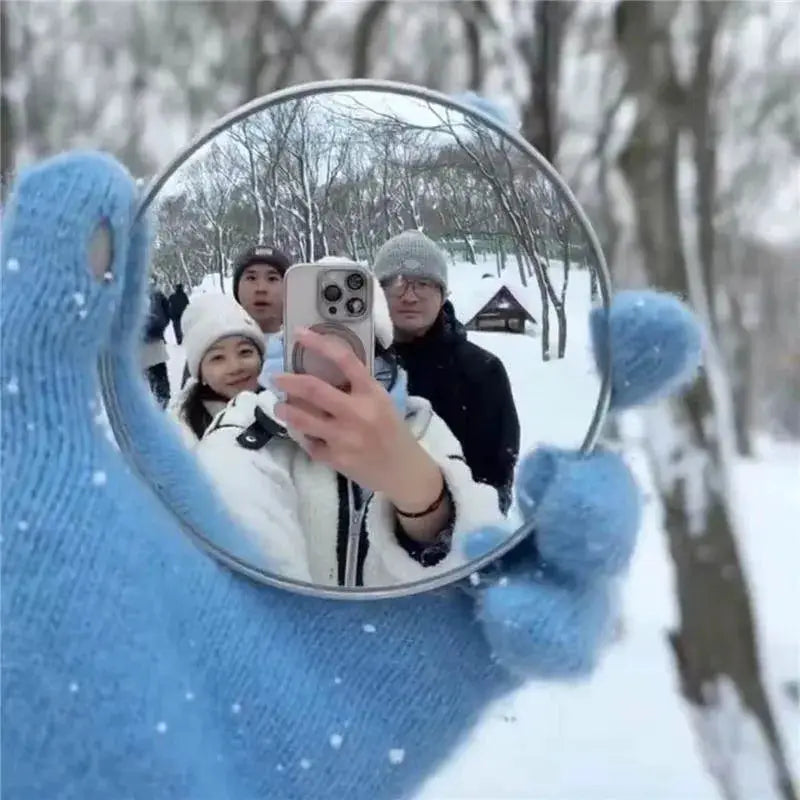 Travel handheld selfie mirror, round convex mirror held by a blue-gloved hand in a snowy forest.