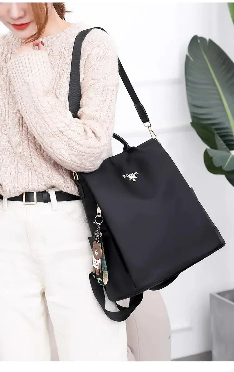 Black Oxford cloth backpack by Simples Market with top handle, adjustable strap, and bear keychain.