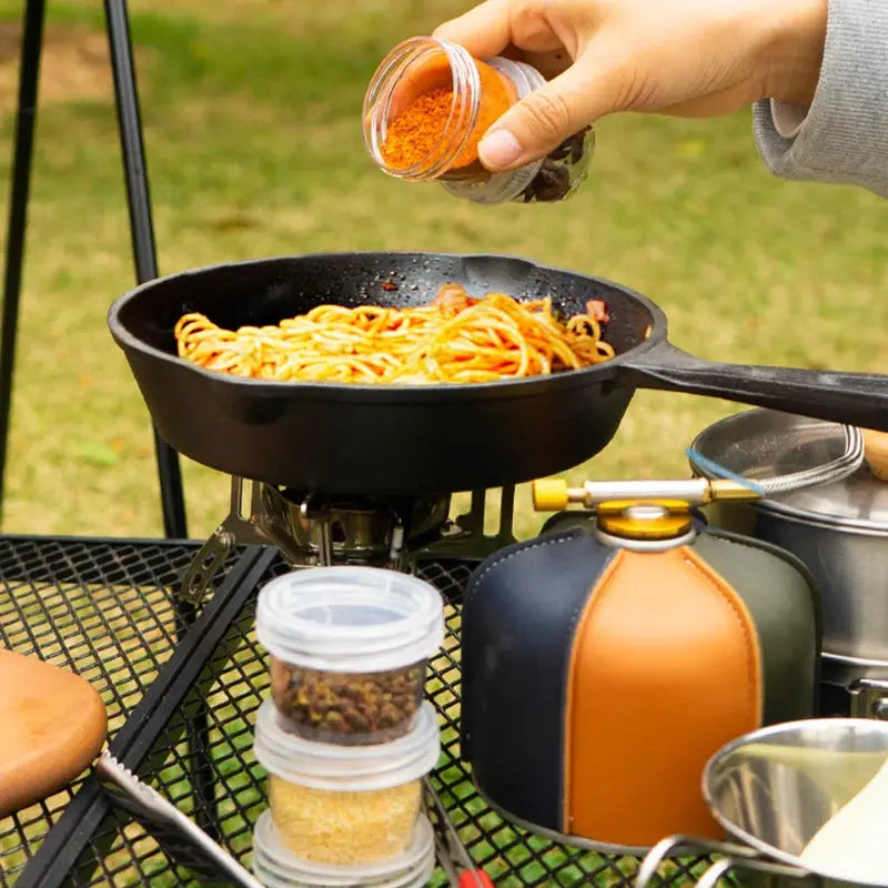 Portable camping spice kit with clear seasoning bottles beside a skillet of pasta on a camp stove
