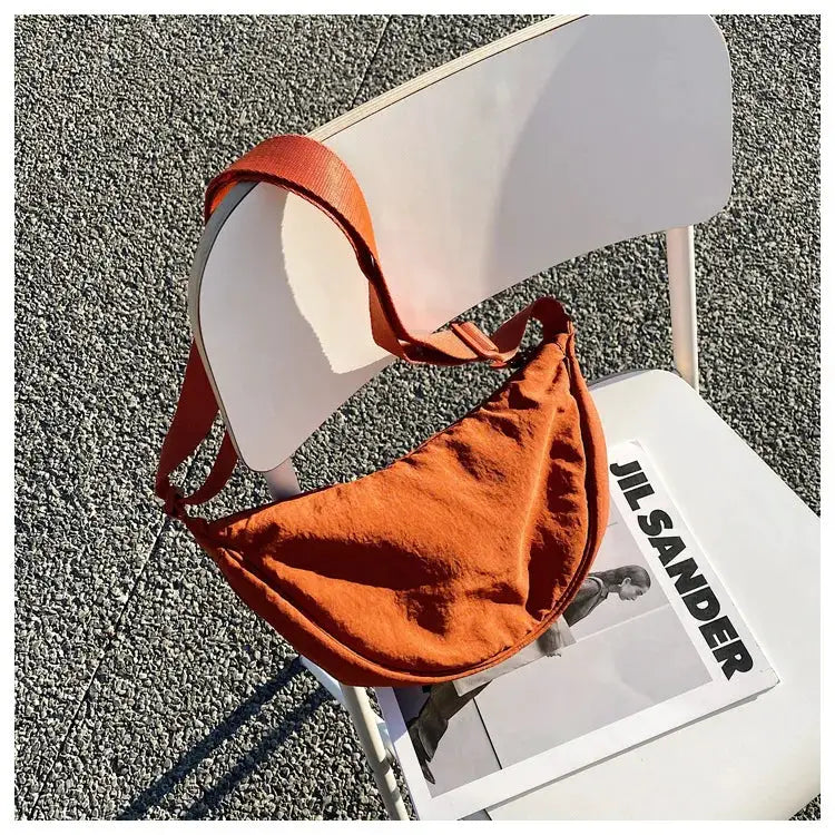 Casual nylon hobo crossbody bag in rust orange with a wide strap, on a white chair beside a Jil Sander magazine.