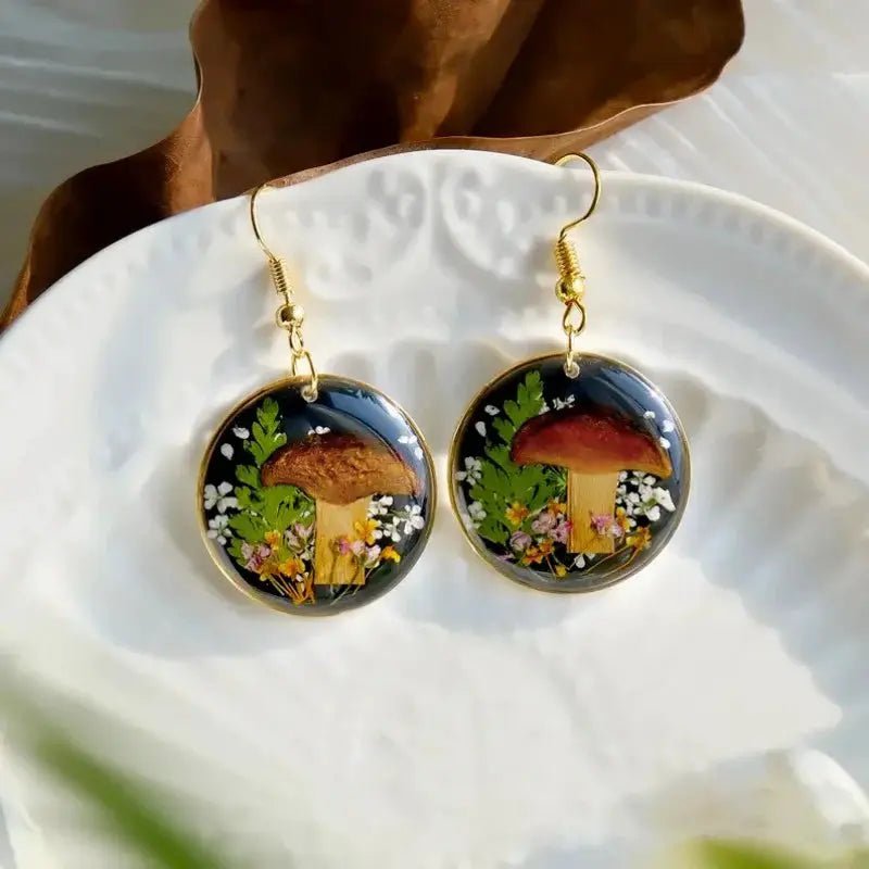 Pair of round epoxy resin earrings featuring pressed mushrooms and wildflowers on black background with gold-tone hooks