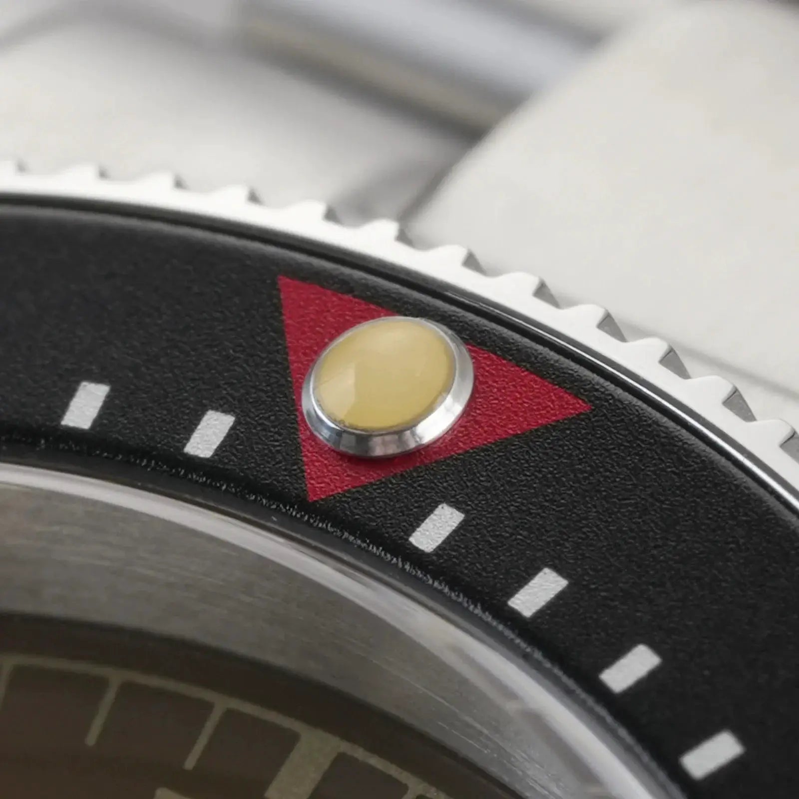 Diver's watch bezel close-up showing black bezel with white teeth, red triangle, and beige luminous dot.