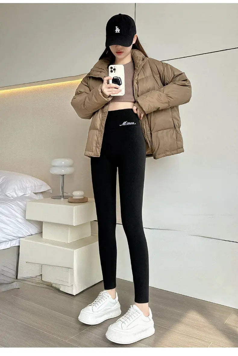 Women's black high-waist leggings with stretchy fit, shown with a beige puffer jacket and white sneakers.