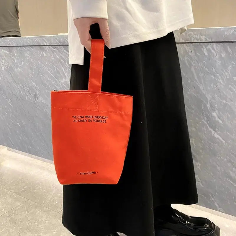 Orange canvas tote bag with short handles, casual shopper style, held by a person in white top and black pants