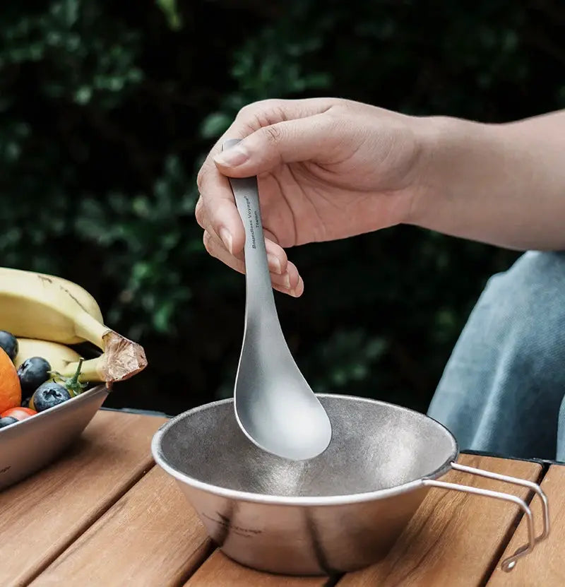 Boundless Voyage titanium camping spoon held over a small metal pot on a wooden table.