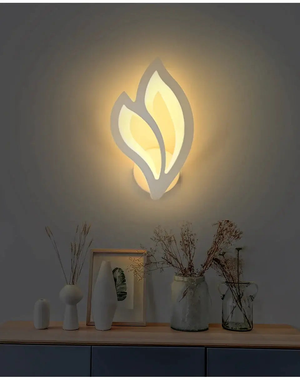 Leaf-shaped modern LED wall lamp in white with warm glow, mounted on wall above a wooden console with vases.