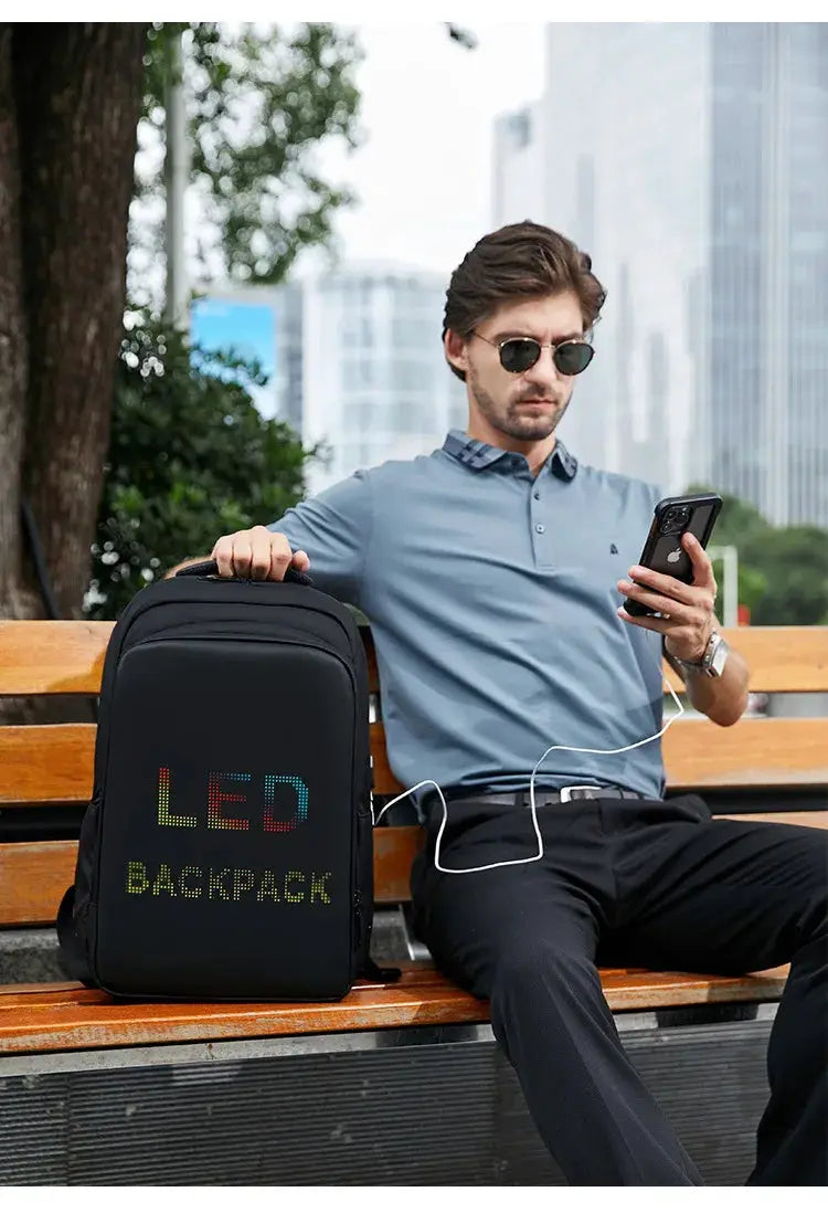 LED display backpack, black with multicolor LED text on the front, shown on a park bench.