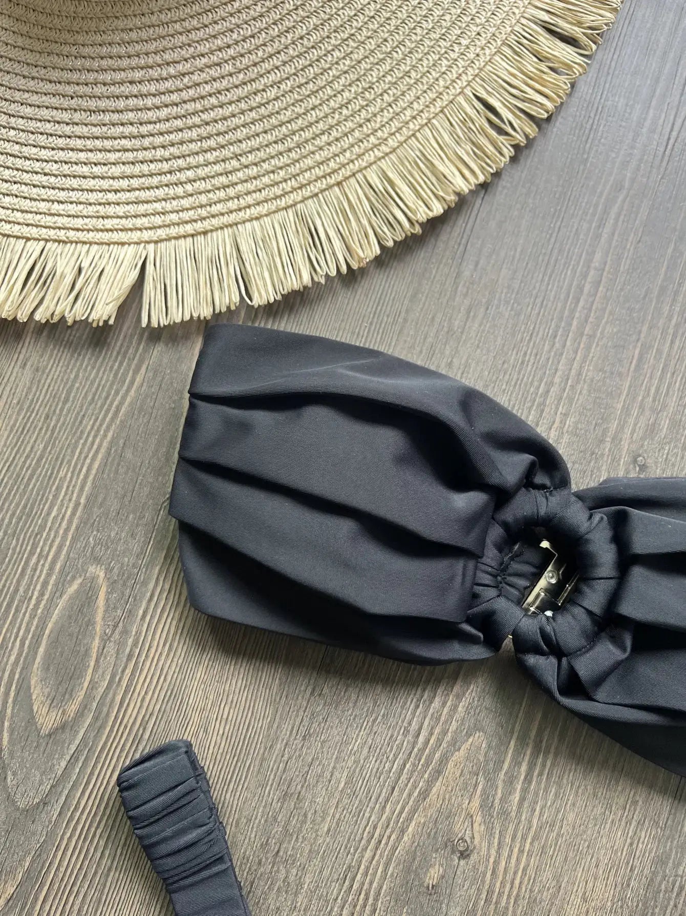 Black micro bikini top and bottom with ruched sides on a wood surface next to a straw hat