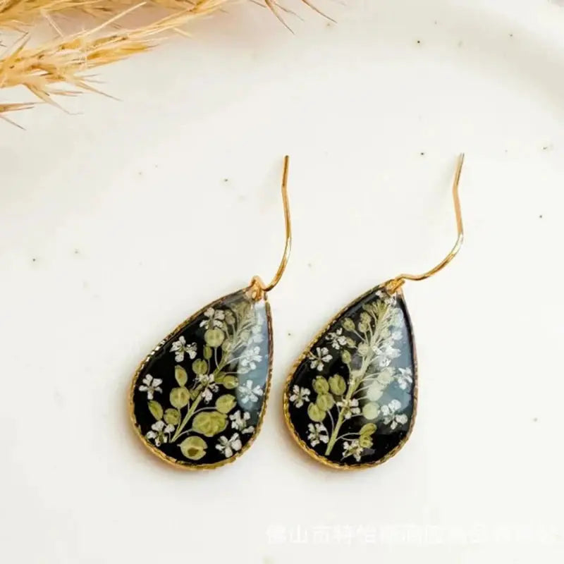 Teardrop pressed-flower epoxy resin earrings with gold trim on black background, green foliage and white blossoms, pair