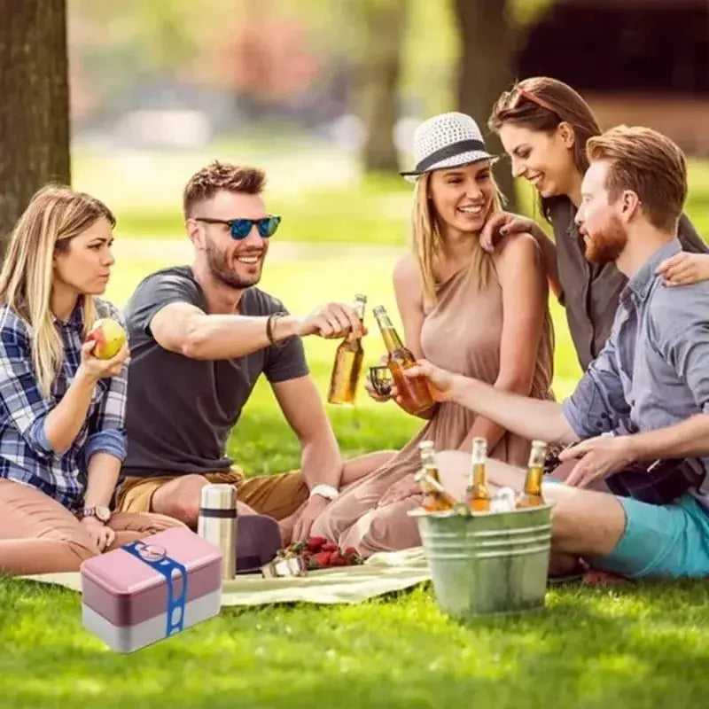 Lunch box in pink and gray sits at a park picnic with friends and bottled drinks.