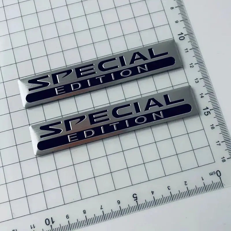 Two 3D metal aluminium car emblems reading SPECIAL EDITION in silver with dark blue lettering