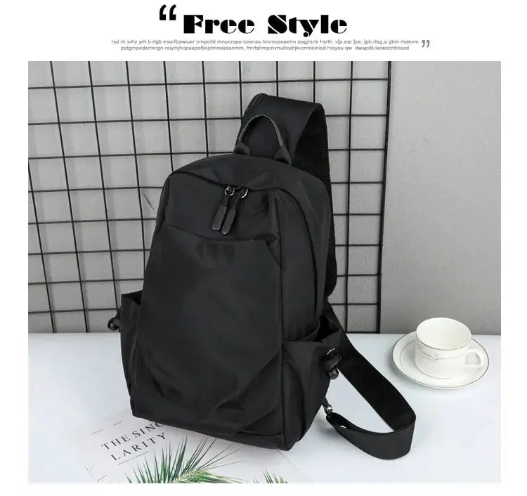 Men's black canvas chest bag with front pocket, double zipper pulls, top handle, and adjustable strap.
