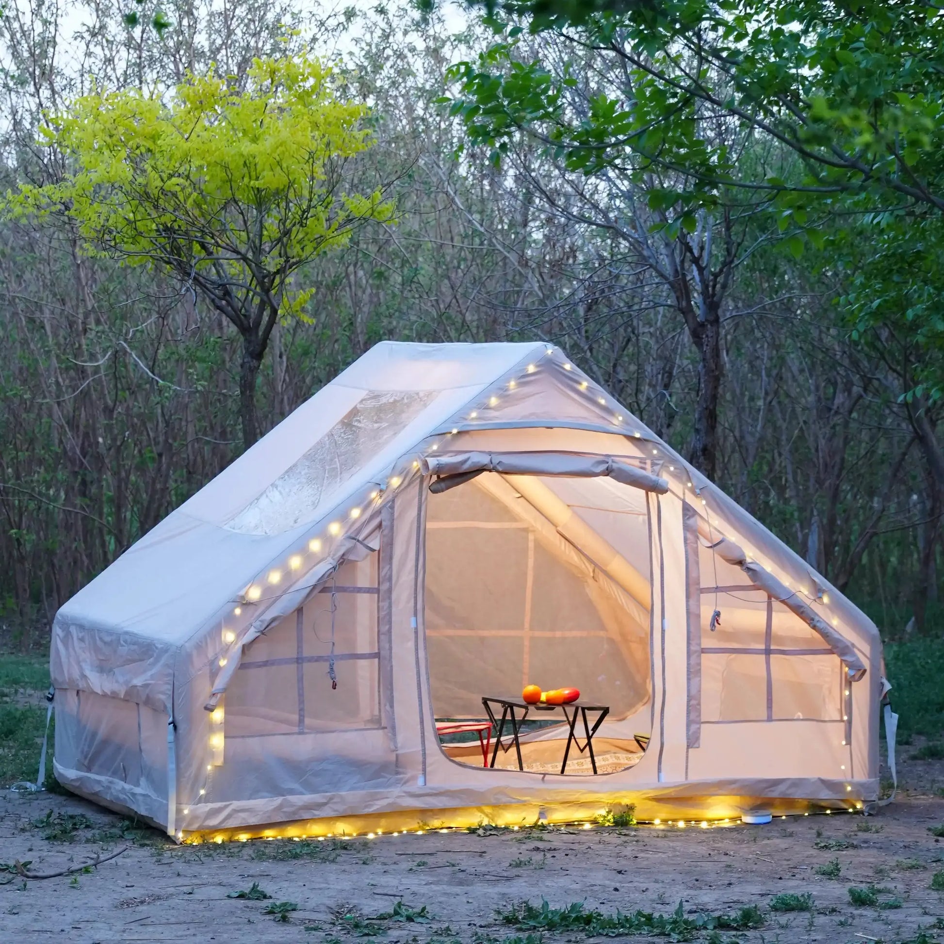 Inflatable camping tent in beige with clear window panels and string lights at a forest campsite.