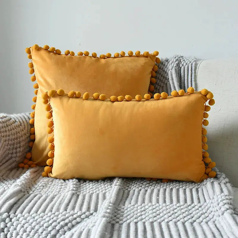 Velvet cushion covers set of 2 in mustard orange with pom-pom fringe, shown on a gray sofa.