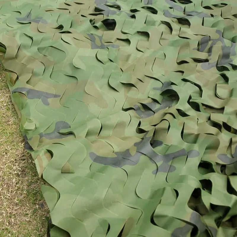 Camouflage netting in woodland pattern with green, tan and brown tones laid over grass.