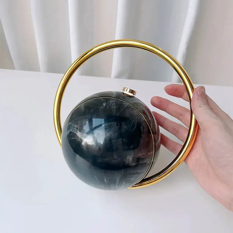 Luxury acrylic evening clutch with gold circular ring handle and dark marble sphere.