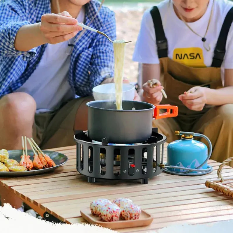 Portable camping stove with black frame and orange handles, pot on top, blue gas canister nearby
