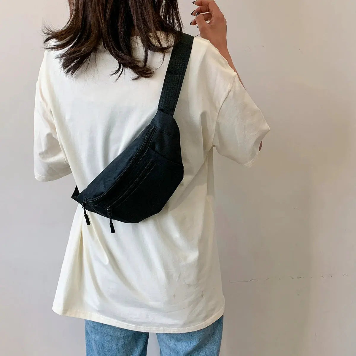 Black crossbody fanny pack with two zip pockets and a wide strap worn by person in a white tee.