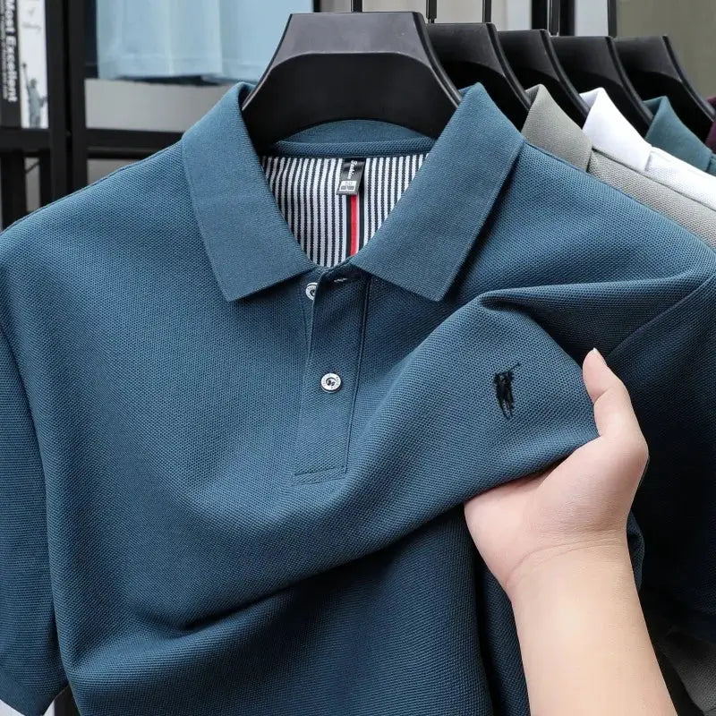 Blue polo shirt on a hanger with small embroidered logo; cotton polo; other polo shirts in various colors visible.