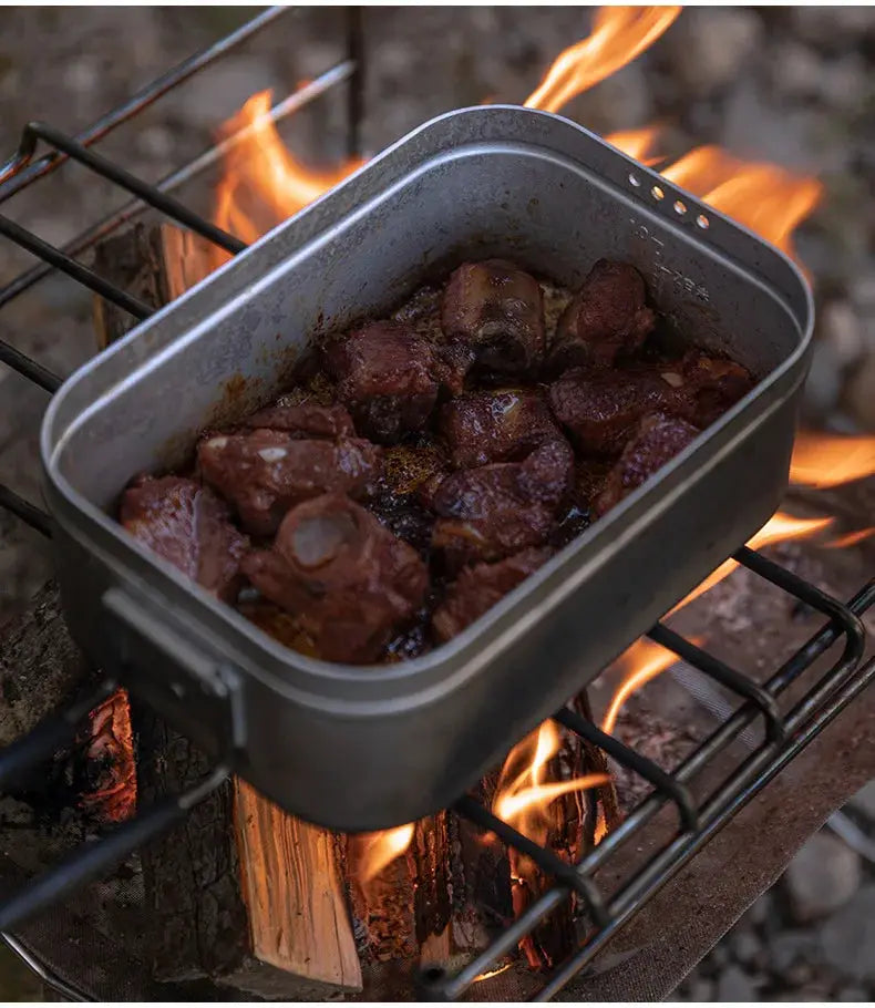 CampingMoon aluminum lunch box on a camp grill over an open flame with browned meat chunks outdoors