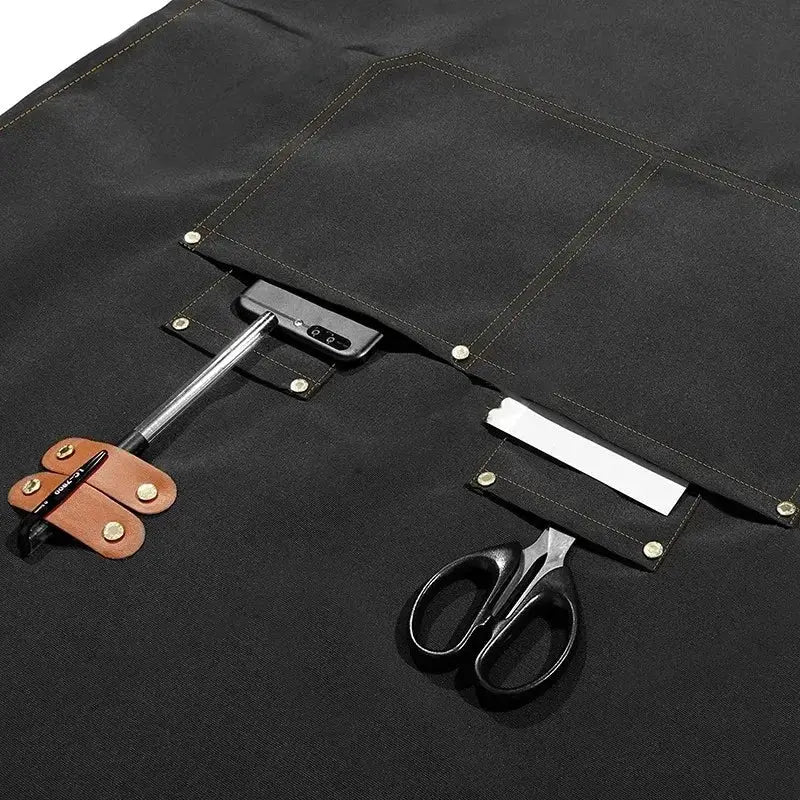 Black waterproof kitchen apron with pockets, scissors and blade tucked in.